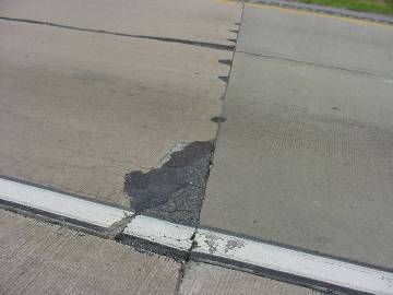 This picture shows a section of a distressed concrete pavement. Severe spalling is observed at the transverse joint and shoulder interface, which was partially repaired with asphalt concrete. Numerous smaller spalls can be seen along the transverse joint.